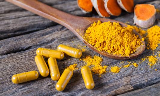 Turmeric powder in wooden spoon, along with turmeric capsules and roots on wood