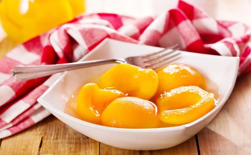 White bowl of halved canned peaches on wooden table, with red and white checkered napkin