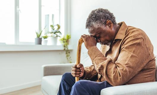 A tired older man sitting in chair, resting his head on his cane